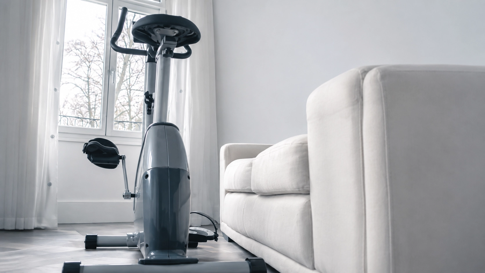 Compact upright exercise bike positioned next to a white sofa in a minimalist living room space