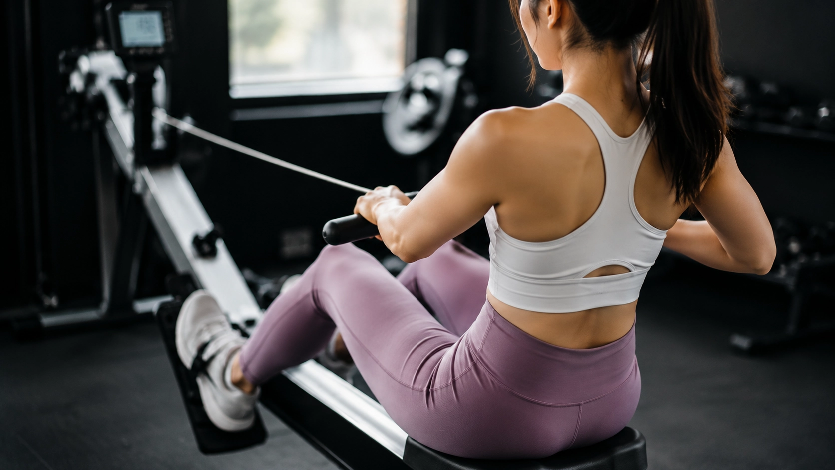 Woman performing rowing machine exercise with proper form, demonstrating full-body conditioning movement for cardiovascular and strength training in a home gym environment