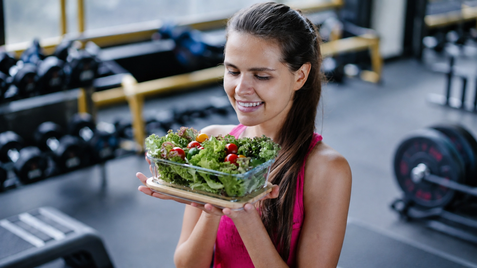 Woman holding fresh vegetable salad in gym setting with dumbbells and weights