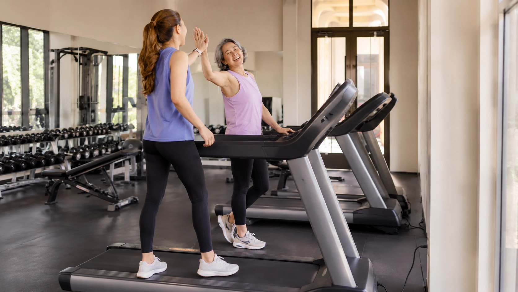 Two women using treadmills with extended handrails in bright gym environment